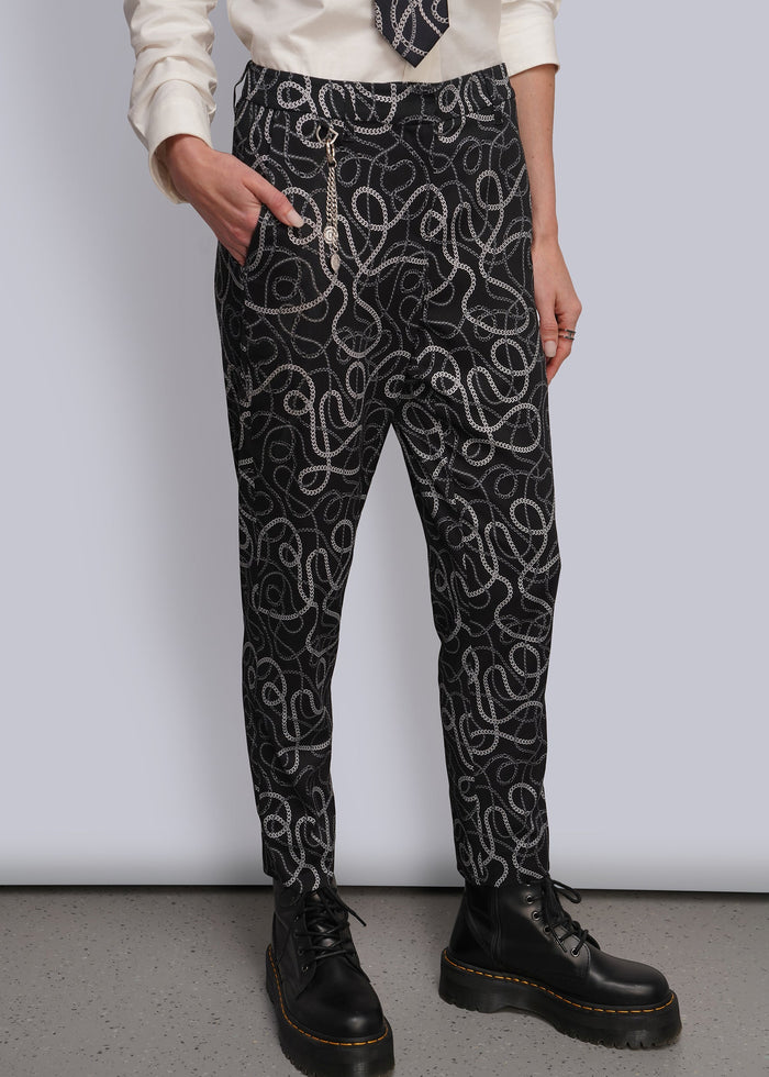 S=A person wearing black pants with a white and grey chain-link wavy pattern, styled with a cream shirt, a patterned tie, and silver chain accessories, paired with black leather boots.