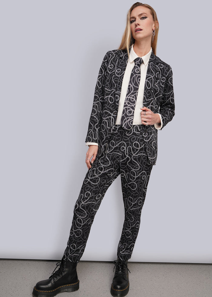 S=A woman modeling a black and white chain-link patterned suit with a white shirt and black boots, standing against a light gray studio background.