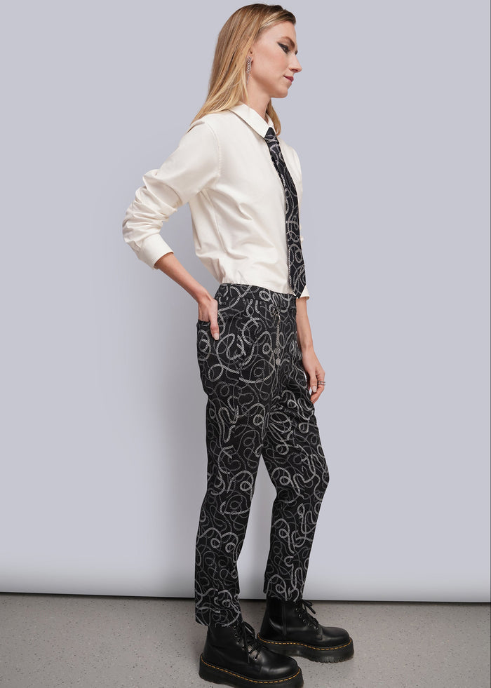 S=A woman stands in profile wearing a cream long-sleeve collared shirt, a black necktie with a chain-link pattern, and black trousers with a matching chain-link print and draped chain details. She is wearing black platform lace-up boots against a light gray studio background.