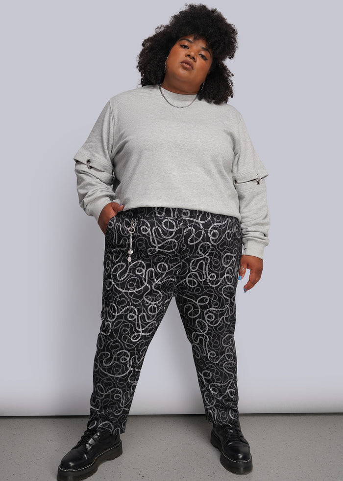 1X=A person modeling a light gray crewneck sweatshirt and black pants with a white and grey chain-link pattern, paired with black boots, against a plain light gray studio background.