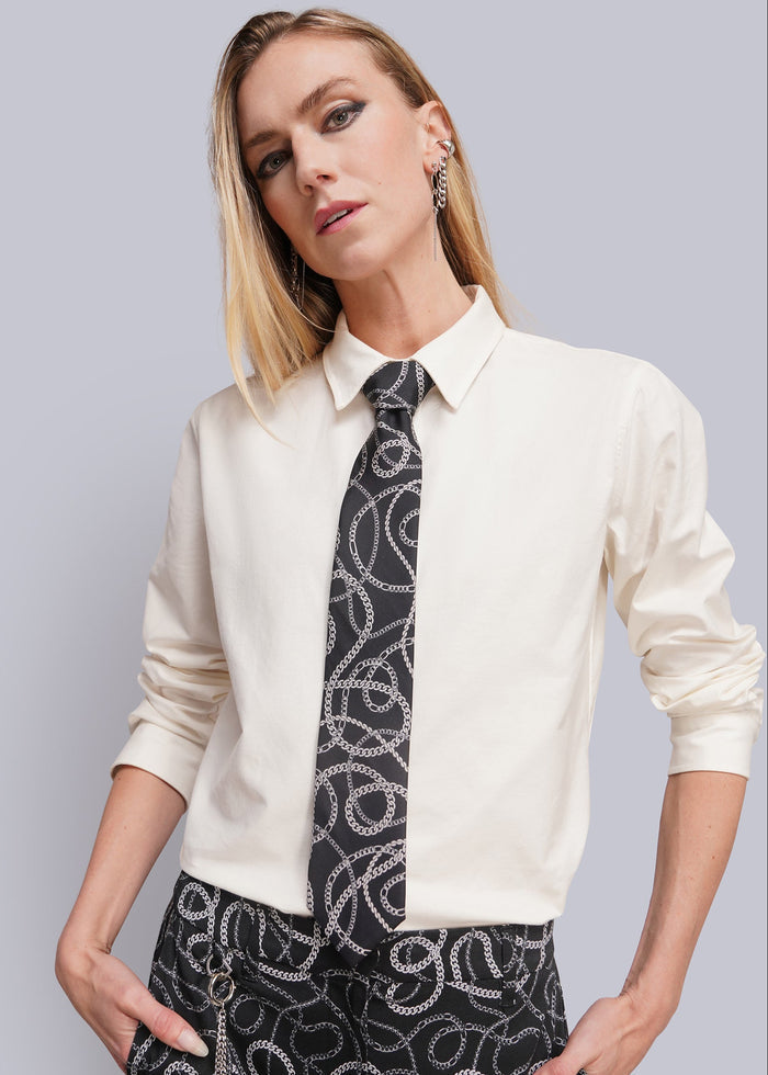 A woman modeling a cream-colored long-sleeve collared shirt paired with a black and white patterned tie and matching patterned trousers, set against a solid light gray background.
