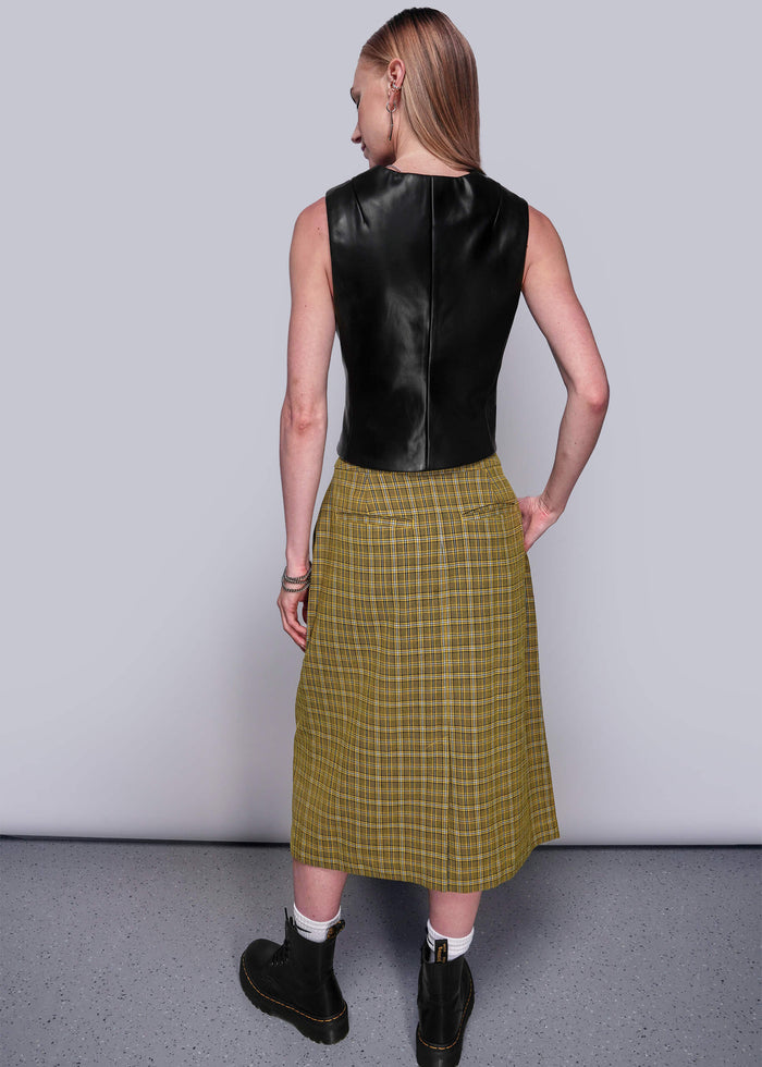 S=Person wearing a black vegan leather vest and yellow plaid skirt against a plain background