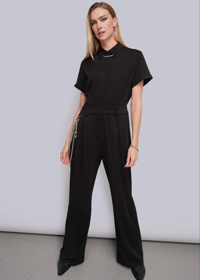 S=A woman modeling a black short-sleeve jumpsuit with a multi chain wallet chain, standing against a light gray studio background.