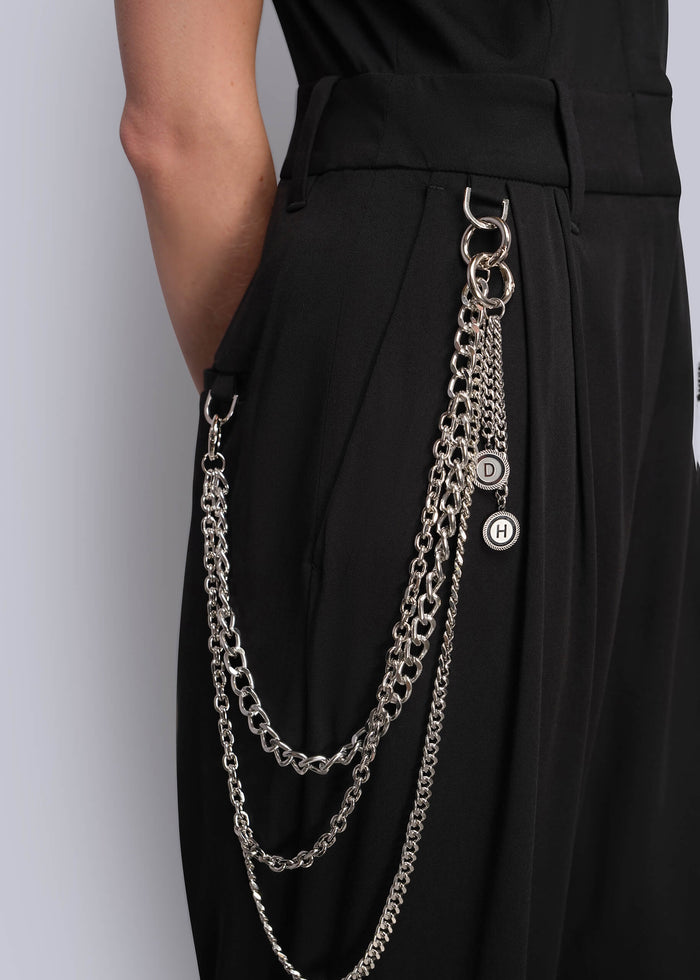 S=A close-up of a person wearing black high-waisted jumpsuit with a long silver wallet chain accessory hanging from the waistband, featuring interlocking circular rings and a small round logo charm.
