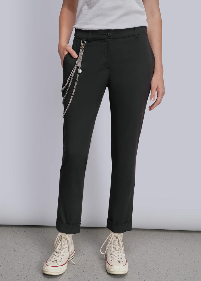 S=A model wearing black slim-fit trousers with a silver chain accessory hanging from the side pocket, styled with a white t-shirt and cream-colored canvas sneakers against a light gray studio background.