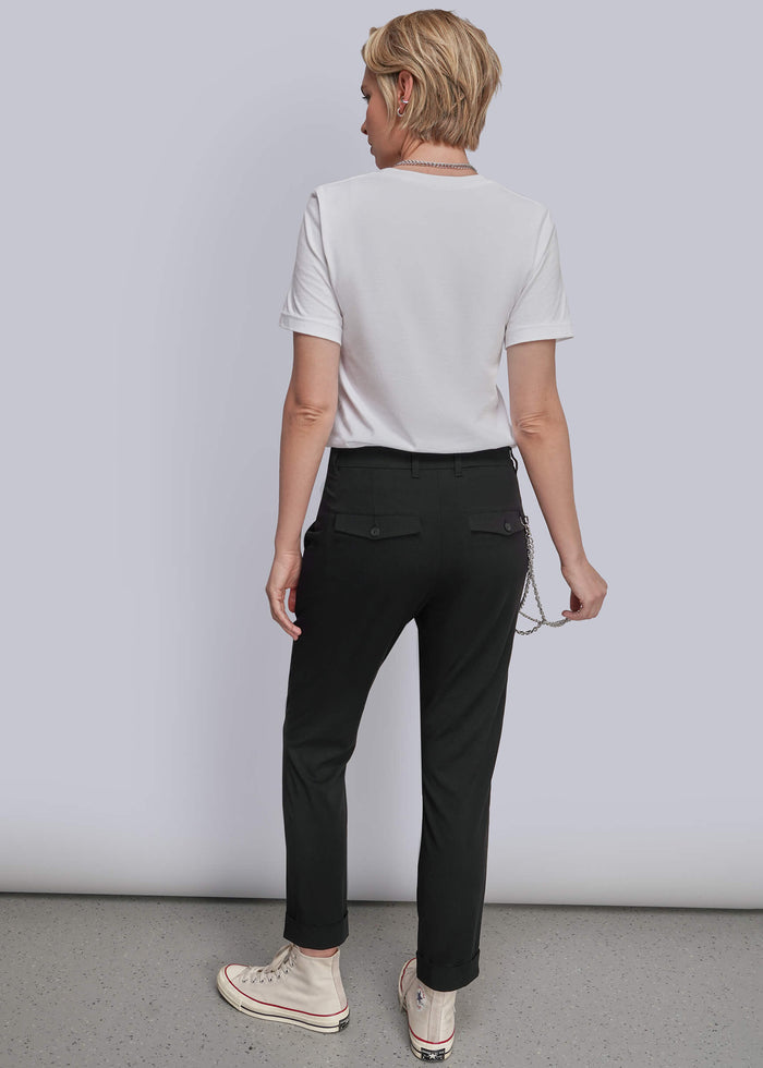 S=A woman shown from the back wearing a white short-sleeve t-shirt, black trousers, and beige sneakers, standing against a light gray studio background.