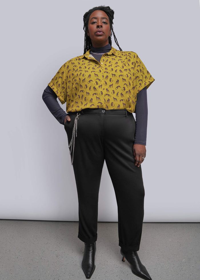 1X=A model stands against a light gray studio wall wearing a yellow short-sleeve button-up shirt with a small dark repeating pattern, layered over a dark gray long-sleeve top, black trousers, and black pointed ankle boots.