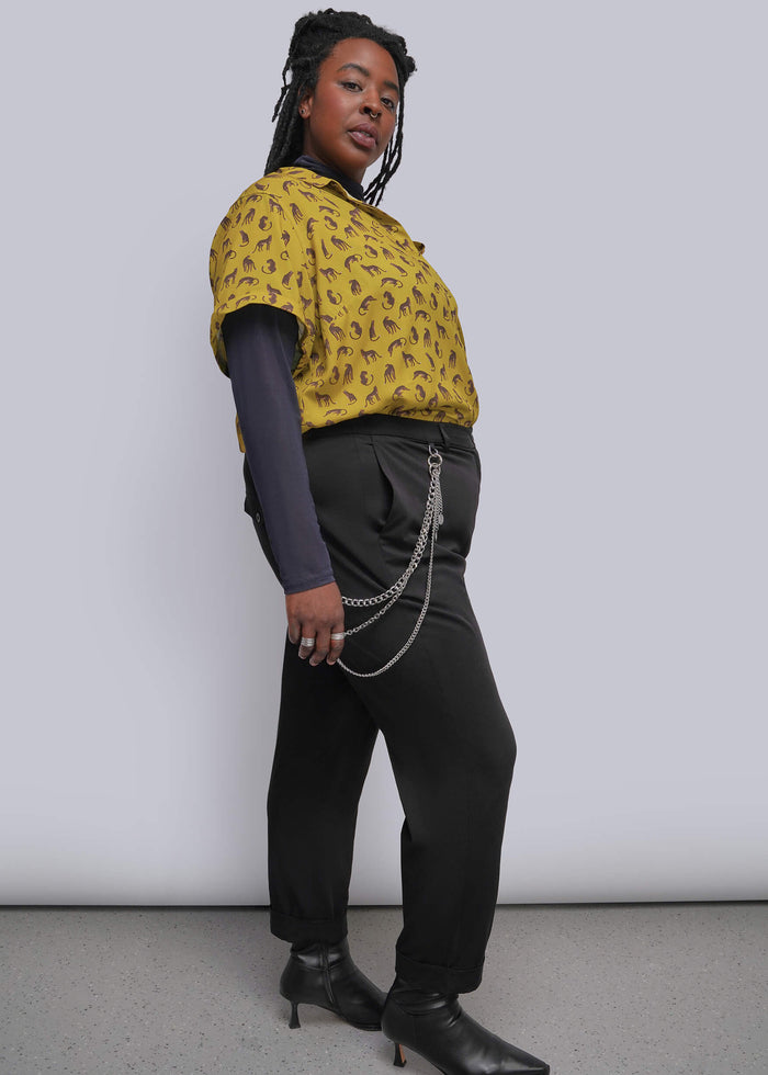 1X=A woman modeling a yellow short-sleeve top with a black animal print, worn over a dark long-sleeve shirt and paired with black pants and black ankle boots, standing against a light gray studio background.