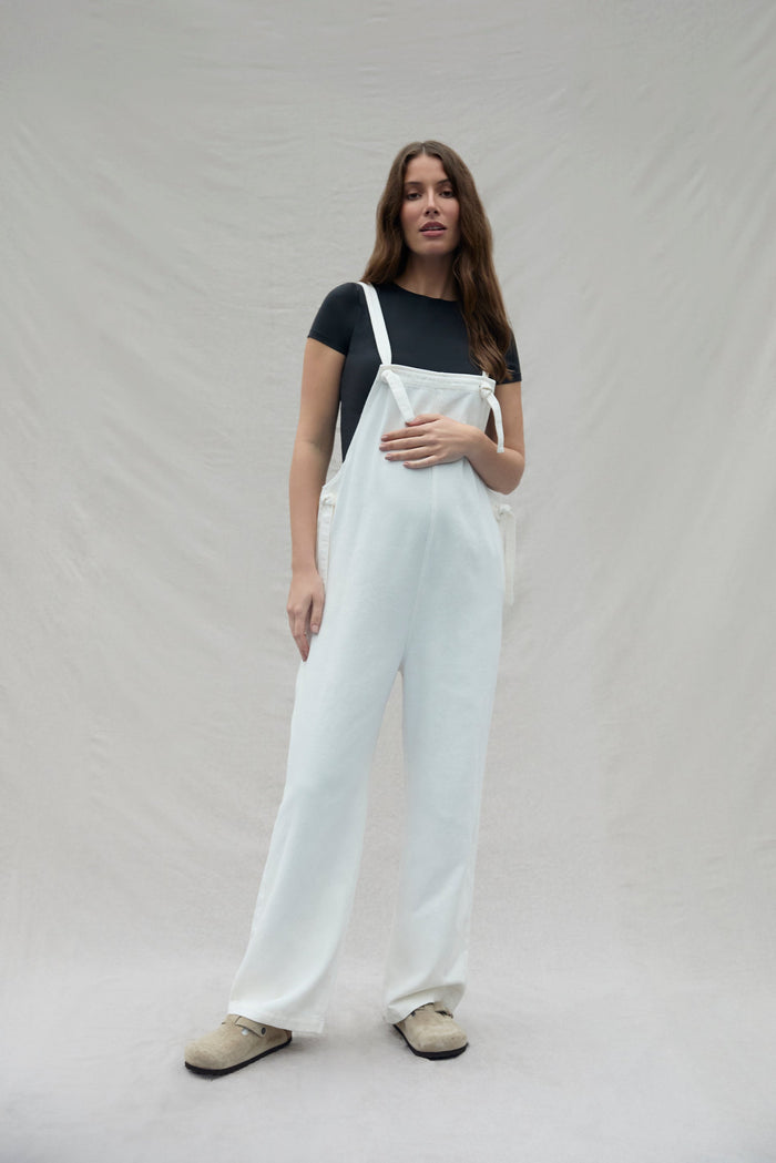 Woman wearing white overalls and a black shirt on a plain background