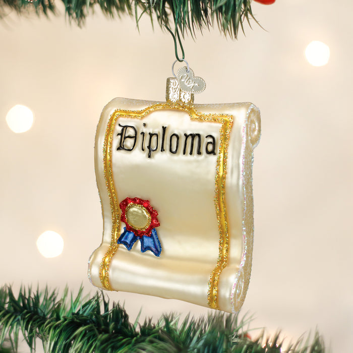 A glass Christmas tree ornament shaped like a rolled diploma, featuring gold glitter edges, the word Diploma, and a red and blue seal, hanging on a tree branch with blurred lights in the background.