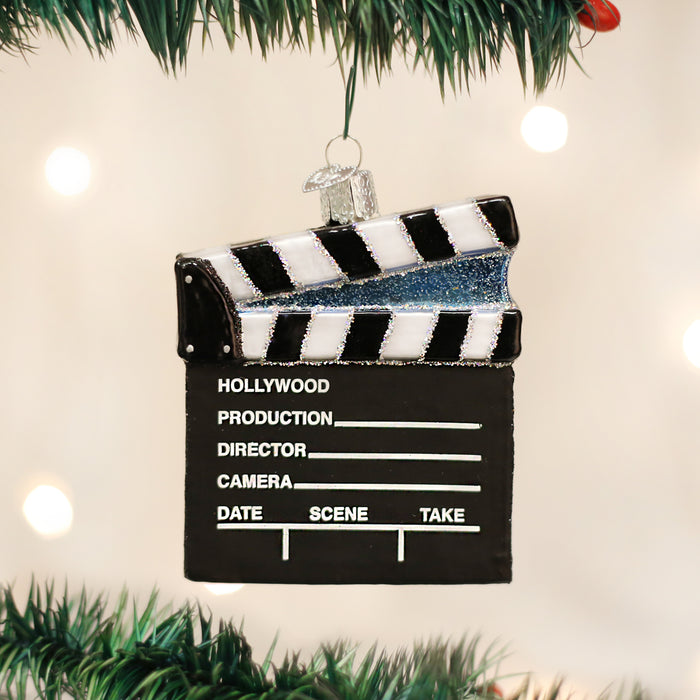 A Christmas tree ornament shaped like a black and white clapperboard with sections labeled Hollywood, Production, Director, Camera, Date, Scene, and Take, hanging among green tree branches.