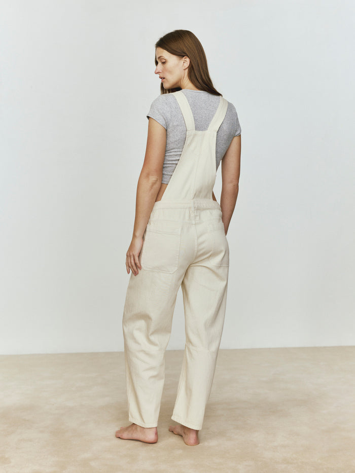 Back view of a woman wearing beige overalls against a plain background