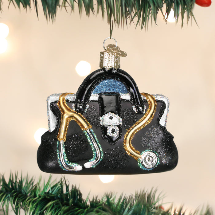 A Christmas tree ornament shaped like a black doctors bag with a gold and silver stethoscope draped over it, hanging from a branch with blurred lights in the background.