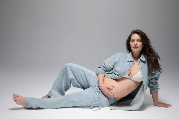 Pregnant woman lying on a gray background wearing light blue denim pants and a denim jacket with a white cotton bra