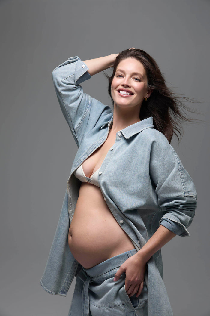 Pregnant woman wearing a light blue denim shirt and pants  with a white cotton bra against a gray background