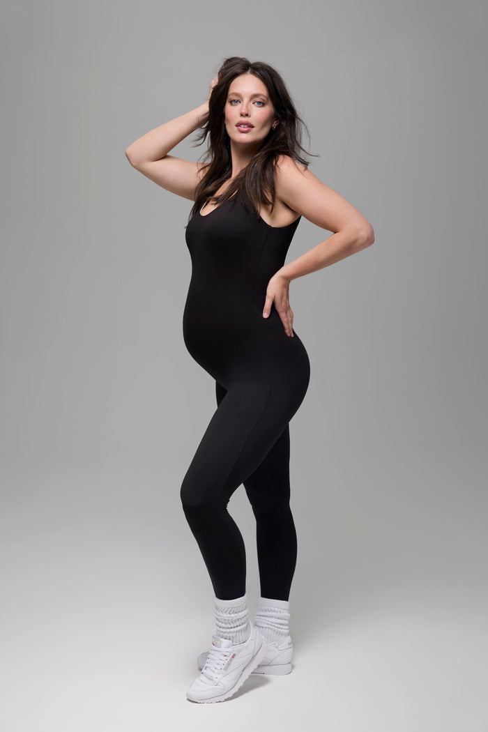 Pregnant woman wearing a black  Bumpsuit bodysuit and white sneakers on a grey background