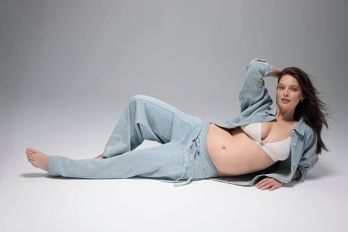 Pregnant woman laying on the floor wearing Bumpsuit denim jeans and button up oversized shirt with a cotton white bra