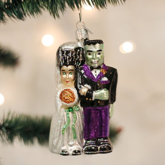 A Christmas tree ornament featuring Frankensteins monster and his bride, dressed in wedding attire with glittery details, hanging from a pine branch against a blurred light background.
