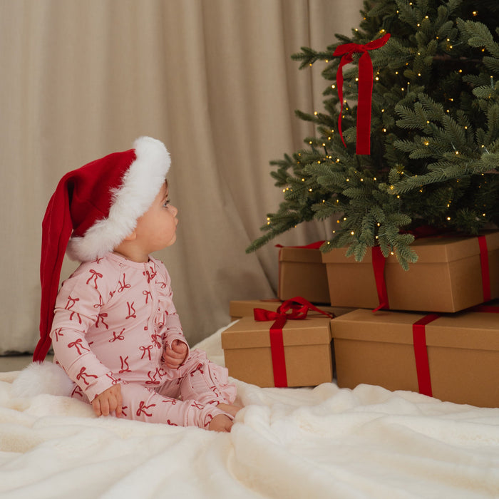 Child in pajamas and Santa hat sitting under a Christmas tree with presents.