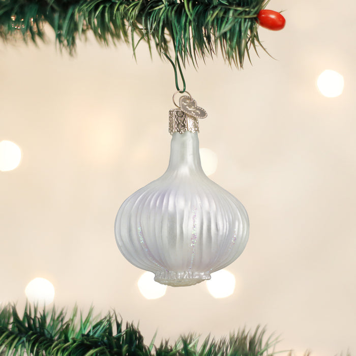 A white, garlic-shaped ornament hangs from a green Christmas tree branch with a silver cap and a small decorative charm, set against a blurred, light background.