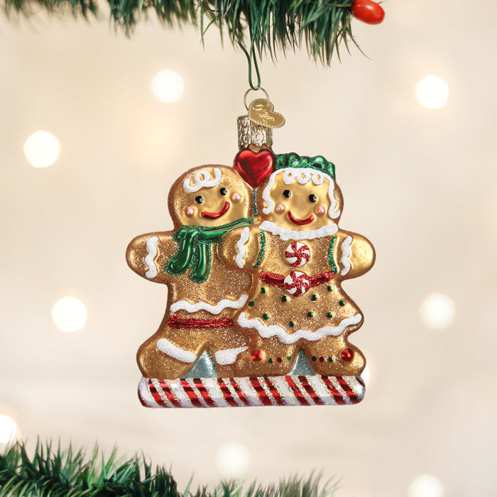 A Christmas tree ornament shaped like two smiling gingerbread people, decorated with icing, scarves, and candy details, hangs from a green branch with blurred white lights in the background.