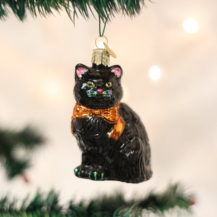 A shiny black cat ornament with green eyes, a gold collar, and pink ears hangs on a Christmas tree with white lights in the background.