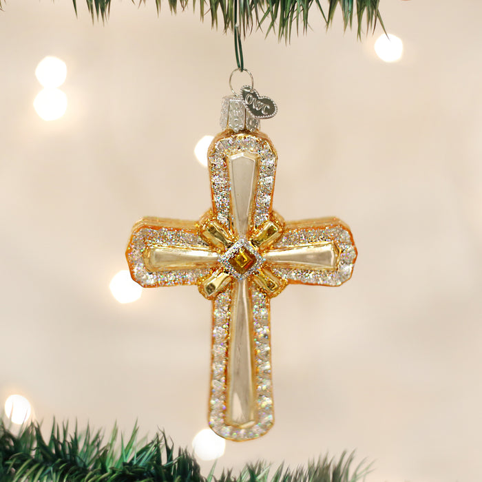 A gold and jeweled cross ornament hangs from a Christmas tree branch, with blurred white lights in the background.