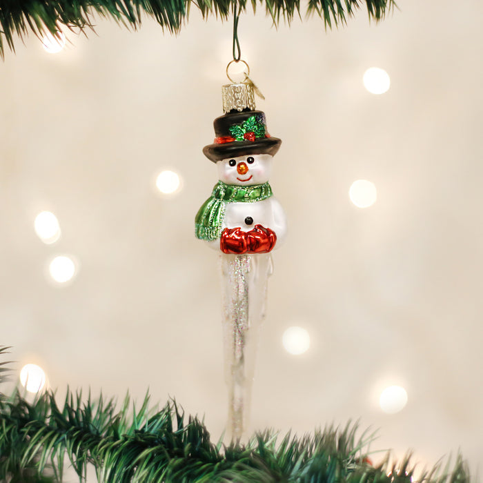 A snowman ornament with a black hat, green scarf, and red mittens hangs on a Christmas tree, surrounded by blurred white lights in the background.