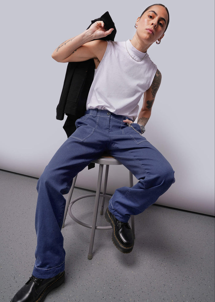 S=Person sitting on a stool wearing a white tank top, blue jeans, and black shoes against a plain background