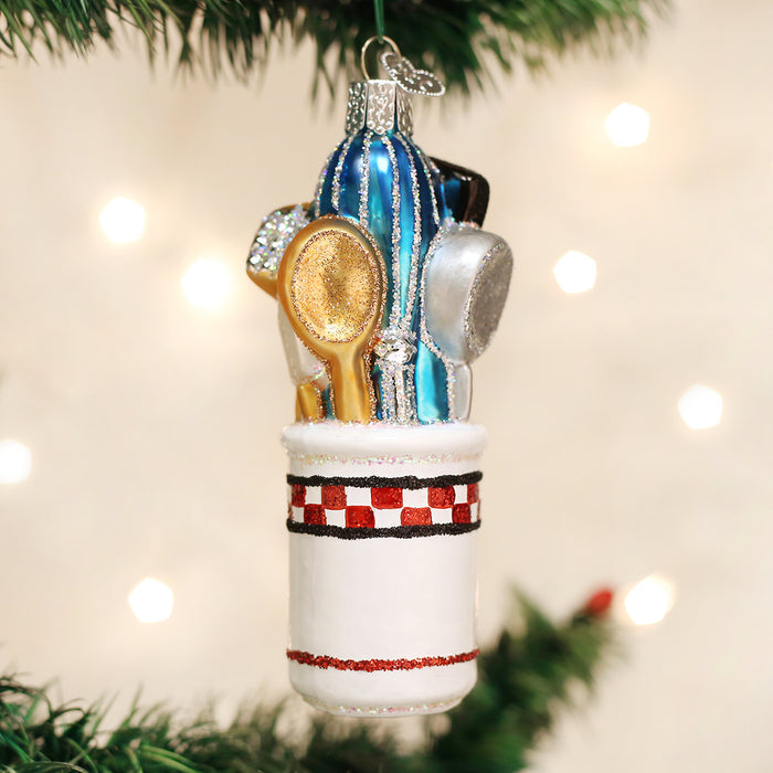 A Christmas tree ornament shaped like a white utensil holder with red checkered trim, filled with miniature kitchen utensils, including a gold spoon, spatula, and blue rolling pin, hanging on a green tree branch.
