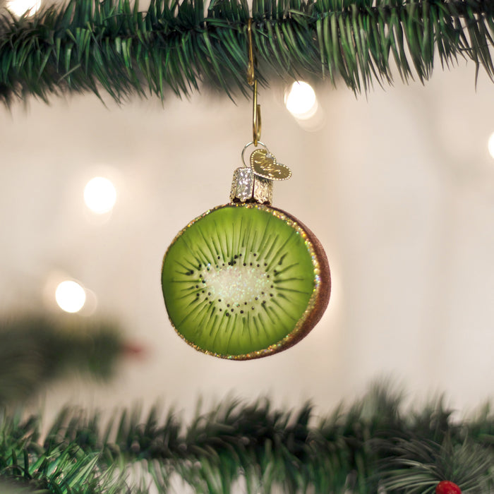 A Christmas tree ornament shaped and painted like a sliced kiwi fruit hangs from a green tree branch, with blurred lights in the background.
