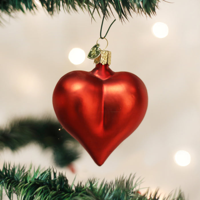 A red, heart-shaped Christmas ornament hangs from a green tree branch, with soft white lights and a blurred background creating a festive atmosphere.