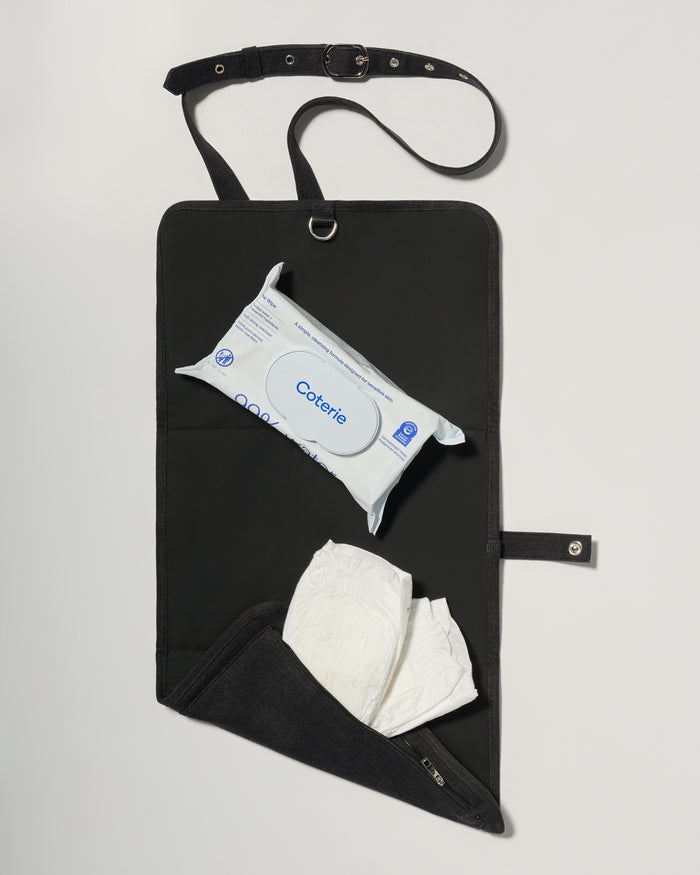 A black fabric diaper bag or organizer pouch with a top strap, shown on a light gray background. The pouch has a front pocket holding a package of baby wipes and a white diaper hanging below.