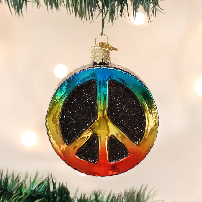 A shiny ornament shaped like a peace sign, decorated in rainbow colors and black glitter, hangs from a Christmas tree branch with a light, blurred background.