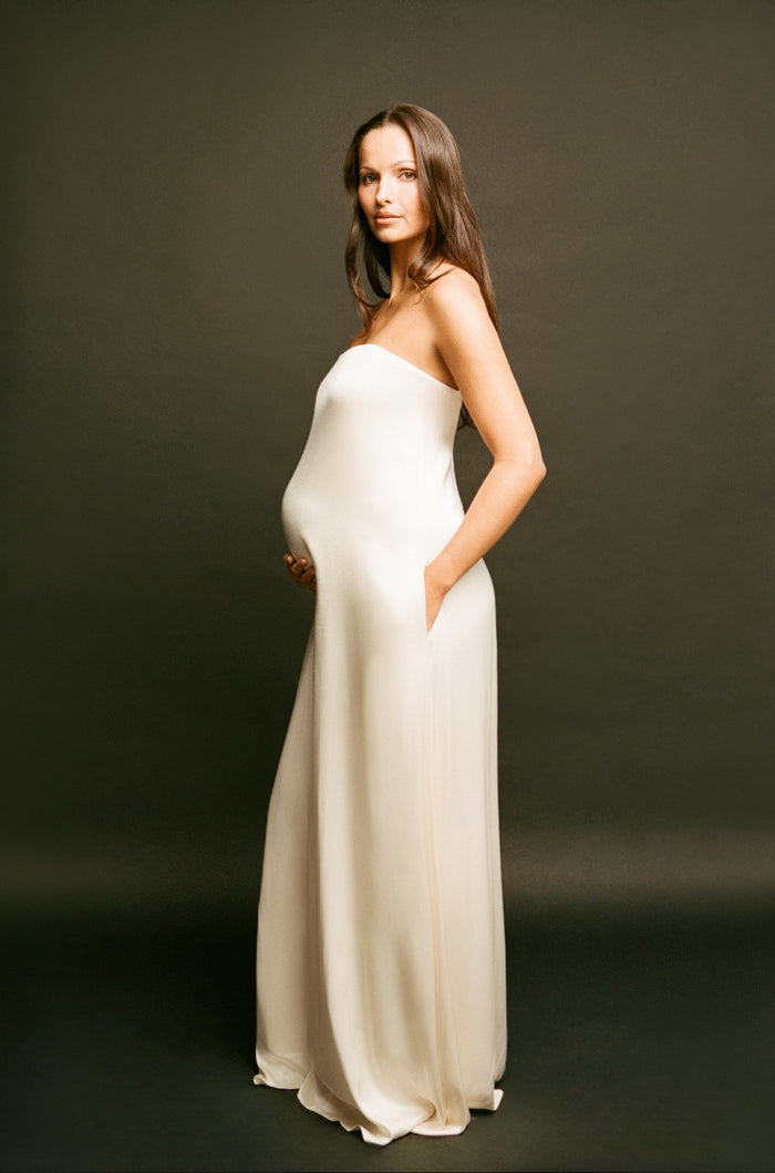 A pregnant woman wearing a Bumsuit strapless, floor-length cream satin gown stands against a dark studio background, holding her baby bump with one hand and resting the other in the side pocket of the dress.