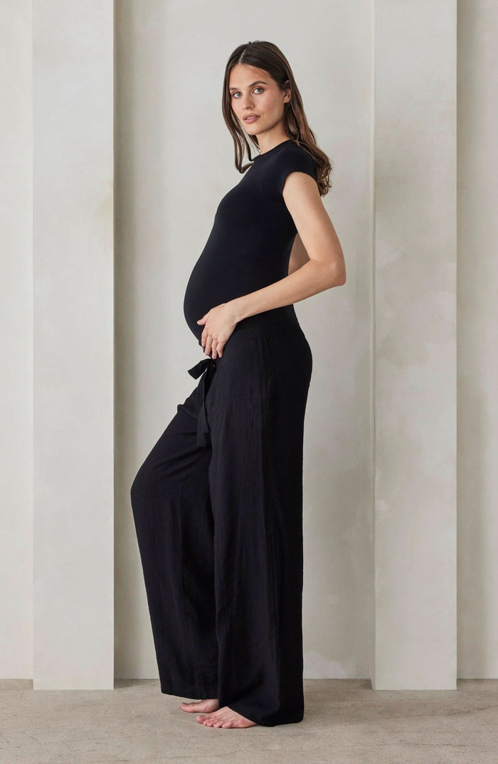 A pregnant woman wearing black relaxed pants and a black shirt by Bumpsuit against a beige feature wall.