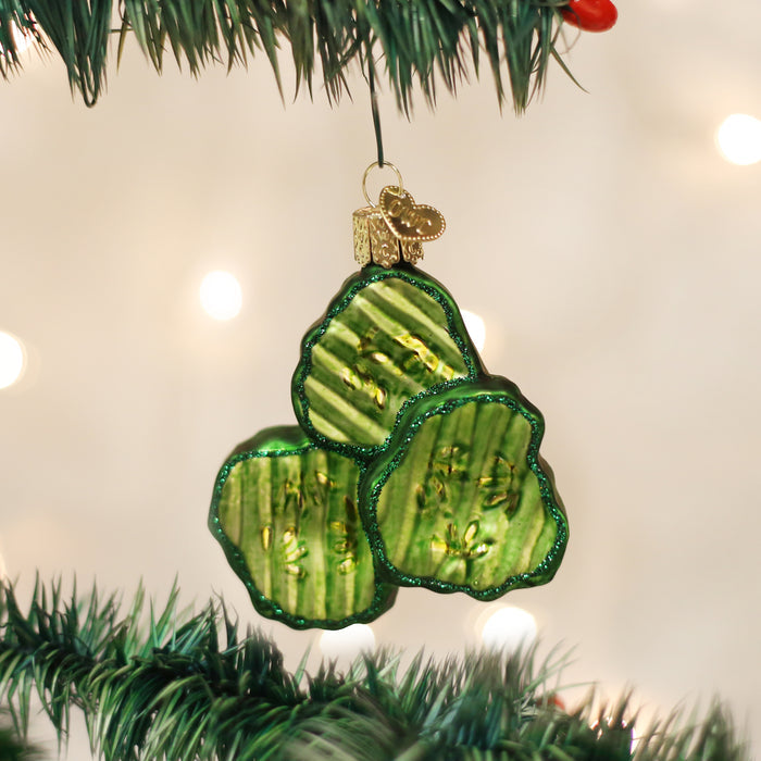 A shiny green glass ornament shaped like three pickle slices hangs from a branch of a Christmas tree, with soft white lights blurred in the background.