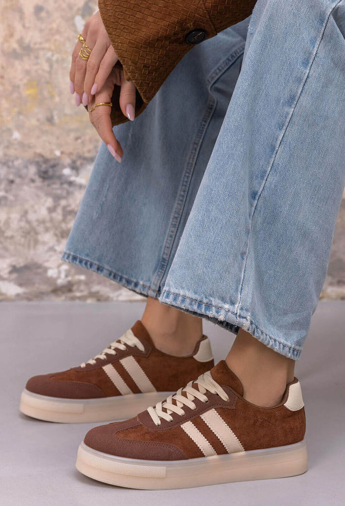 A person wearing brown suede and canvas low-top sneakers with cream stripes and laces, paired with light-wash wide-leg jeans and gold rings, seated against a textured wall.