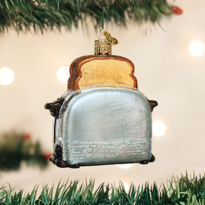 A glittery toaster-shaped ornament with two slices of toast hangs on a green Christmas tree branch, with soft white lights in the background.
