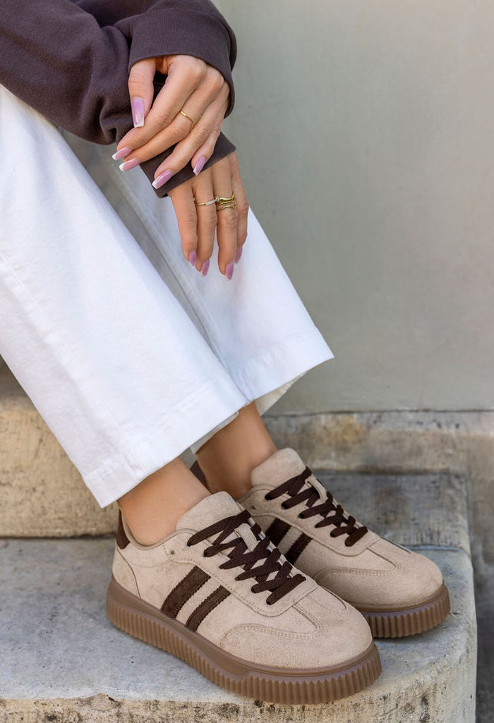 A person wearing beige low-top sneakers with dark brown laces and three dark brown stripes on the side, paired with white pants and a dark brown top, sitting on stone steps.