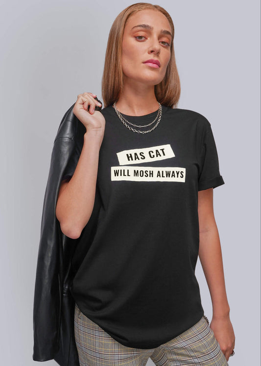 S=A woman modeling a black graphic t-shirt with the text 'HAS CAT WILL MOSH ALWAYS' in white block letters. She is wearing plaid pants and holding a black leather jacket over her shoulder, set against a plain gray studio background.