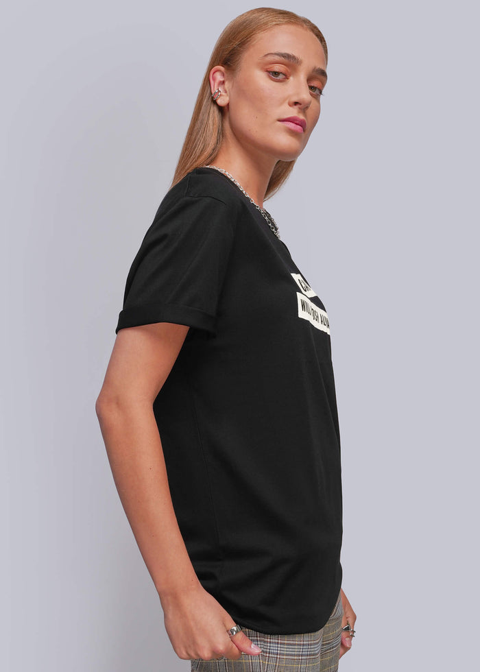 S=A woman wearing a black short-sleeve graphic t-shirt with a white printed design on the chest, paired with plaid trousers, photographed against a solid light gray studio background.