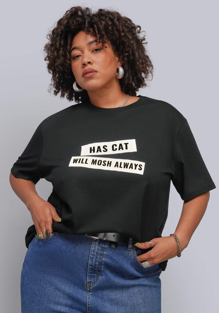 1X=A woman wearing a black graphic t-shirt with the text 'HAS CAT WILL MOSH ALWAYS' in white block letters, paired with blue jeans, against a solid gray studio background.