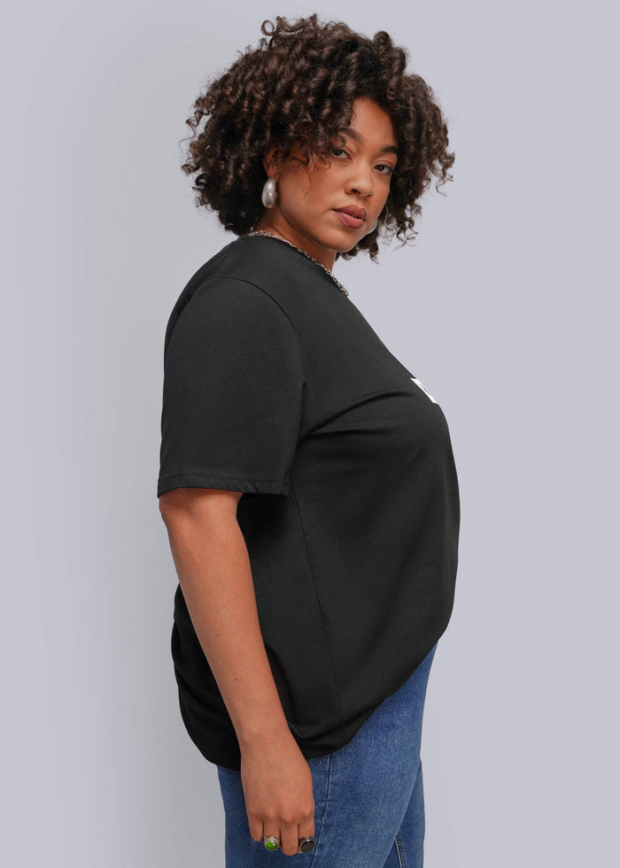 1X=A woman with curly dark hair is shown from a side-back angle wearing a loose-fitting black short-sleeve t-shirt and blue jeans against a solid light gray background.