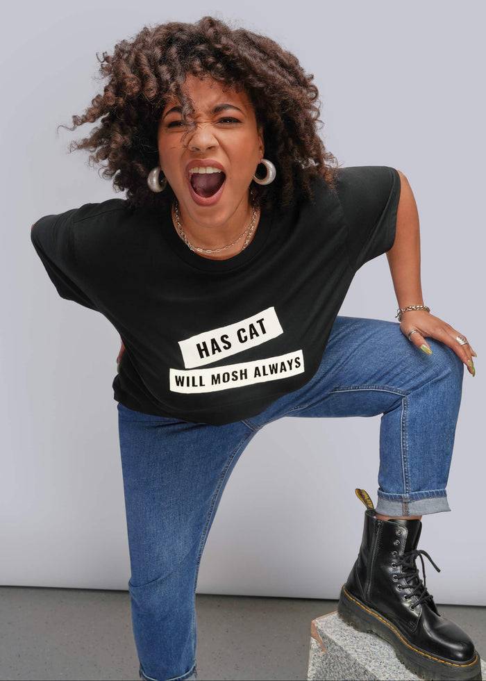 1X=A woman posing in a black graphic t-shirt with the text 'HAS CAT WILL MOSH ALWAYS' in white, paired with blue jeans and a black lace-up boot, set against a plain light gray studio background.