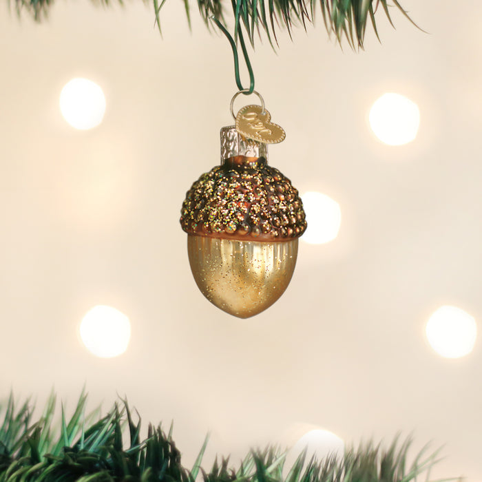 A gold acorn-shaped Christmas ornament with a glittery cap hangs from a green tree branch, with soft white lights glowing in the blurred background.