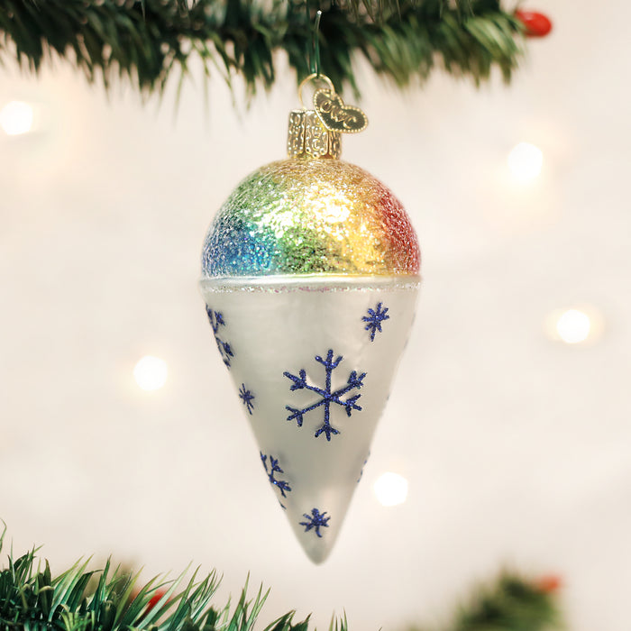 A Christmas tree ornament shaped like an ice cream cone, with a glittery rainbow top and white base decorated with blue snowflakes, hangs from a green tree branch.