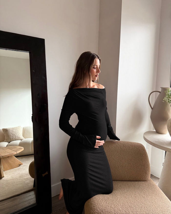 A pregnant woman wearing a Bumpsuit fitted black off-the-shoulder maternity dress, standing next to a beige textured armchair in a bright, modern room with a large dark-framed mirror.