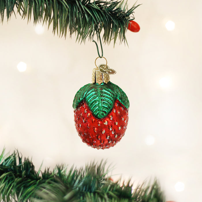 A strawberry-shaped ornament hangs from a green Christmas tree branch, with a soft, white, out-of-focus background and twinkling lights.