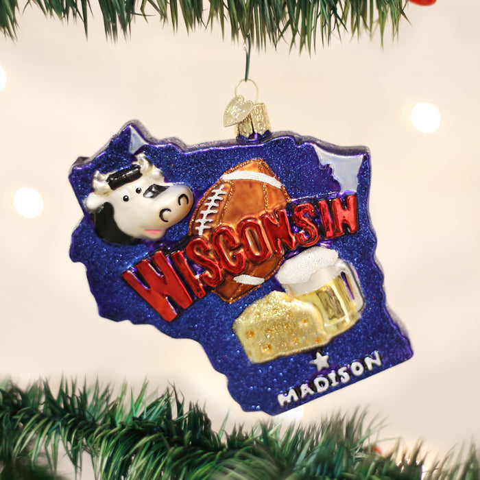 A Christmas ornament shaped like the state of Wisconsin features a cow, a football, a mug of beer, and the words WISCONSIN and MADISON in red and white letters, hanging from a green tree branch.